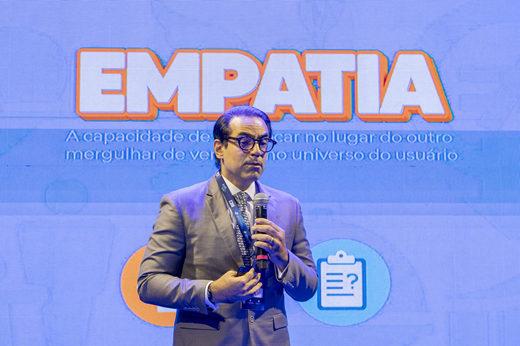Juiz Julio Coelho fala ao microfone na abertura do evento. Ele traja terno cinza. Atrás dele, está um telão azul onde se lê a palavra "empatia".
