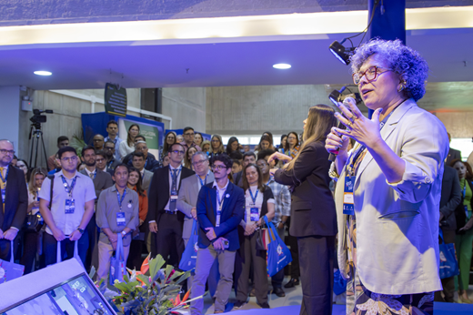 A vice-reitora Diana Azevedo fala ao microfone na abertura do evento. De lado no palco, ela traja blazer verde-claro. No fundo da imagem, vê-se o público, também em pé.