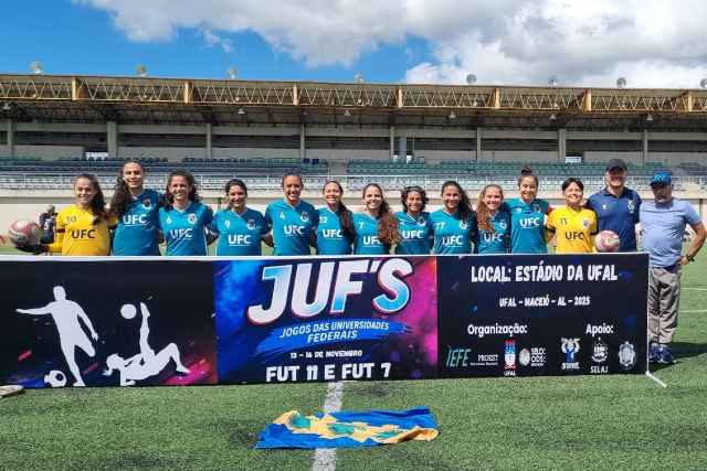 Imagem: Foto de um time feminino de futebol da Universidade Federal do Ceará posando em campo aberto, sob céu azul com algumas nuvens. As atletas estão alinhadas atrás de uma faixa grande e colorida dos JUF’s – Jogos das Universidades Federais, que traz ilustrações de jogadores em ação e informações sobre o evento. O grupo é composto por 14 jogadoras vestindo uniformes azul-turquesa com o nome “UFC” no peito, além de dois membros da comissão técnica nas extremidades, ambos usando boné; um deles veste camisa azul-marinho e o outro camisa azul clara. Algumas atletas seguram bolas de futebol. A faixa à frente destaca os dizeres “Fut 11 e Fut 7”, o local do evento — Estádio da UFAL, em Maceió (AL) — e logotipos de organizações e apoios. No gramado, em frente à faixa, há uma bandeira azul e amarela estendida.