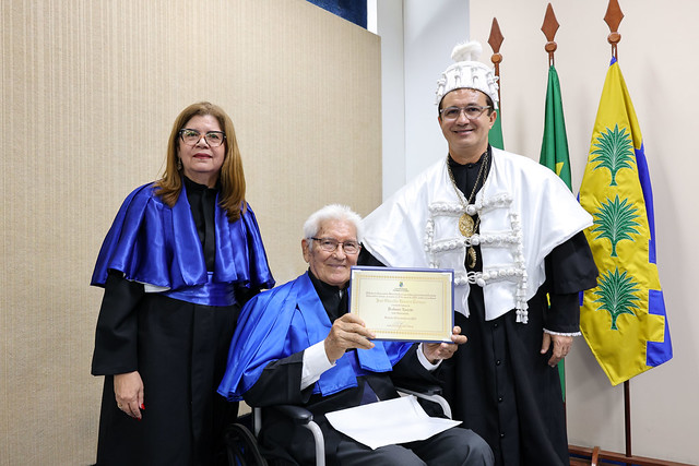 O professor José Osvaldo Beserra Carioca, ao lado do reitor Custódio Almeida e da diretora do CCA, Sônia de Oliveira: noite para recordar e celebrar conquistas (Foto: Viktor Braga/UFC) Imagem: Uma mulher com beca azul e um homem com traje cerimonial branco posam ao lado de uma pessoa idosa sentada em cadeira de rodas, que segura um certificado. As bandeiras oficiais estão posicionadas atrás deles.