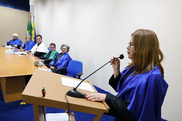 A professora e diretora do CCA, Sônia Maria de Oliveira: título concedido expressa a "gratidão profunda da UFC e de todos que encontram inspiração nesses professores" (Foto: Viktor Braga/UFC) Imagem: Mulher usando beca azul fala ao microfone em um púlpito de madeira, segurando um papel. À sua frente, uma mesa comprida onde várias pessoas, também com becas coloridas, estão sentadas observando. Ao fundo, bandeiras oficiais estão posicionadas no canto da sala.