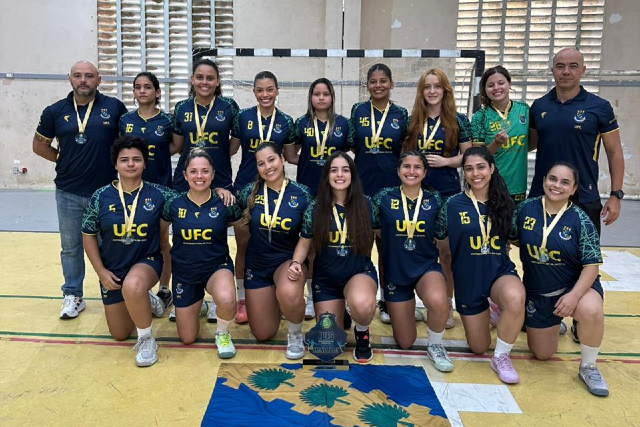 A seleção feminina da UFC conquistou o título de tetracampeã nos Jogos das Universidades Federais (JUFs) de Handebol 2025 (Foto: Divulgação) Imagem: Fotografia de um time feminino de handebol da Universidade Federal do Ceará (UFC). Quinze pessoas posam em quadra: treze atletas e dois treinadores, distribuídos em duas fileiras. Na fileira da frente, sete jogadoras estão agachadas ou ajoelhadas, todas usando uniforme azul-marinho com detalhes em verde e amarelo, o brasão da UFC e seus respectivos números. Na fileira de trás, seis jogadoras e dois treinadores estão em pé. Todas as atletas usam medalhas douradas no pescoço e sorriem para a foto. No chão, à frente da equipe, há uma bandeira azul com desenhos em verde e amarelo. Ao fundo, vê-se a trave do gol e paredes claras com janelas altas que deixam entrar luz natural.