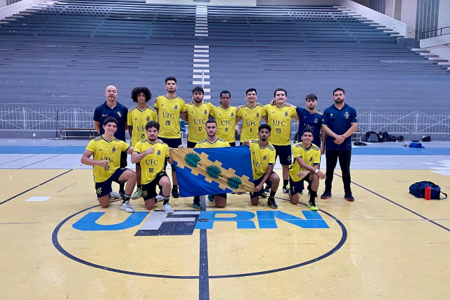 Os bicampeões ficaram em quarto lugar na classificação final (Foto: Divulgação) Imagem: Fotografia de um time masculino de handebol da Universidade Federal do Ceará (UFC). O grupo está posicionado no centro de uma quadra esportiva, sobre o logo pintado no chão da UERN. São treze pessoas ao todo: onze atletas e dois integrantes da comissão técnica. Na fileira da frente, cinco jogadores estão agachados segurando, ao centro, uma bandeira azul com desenhos em verde e amarelo. Na fileira de trás, seis atletas estão em pé, todos vestindo uniforme amarelo com detalhes em azul-marinho e o nome “UFC” no peito. À esquerda e à direita, dois treinadores usam camisa azul-marinho e posam ao lado do grupo. As arquibancadas ao fundo estão vazias e o ginásio é amplo e bem iluminado.