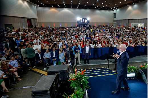 Imagem: Trajando terno azul marinho, o presidente Lula discursa em um palco do Centro de Eventos do Ceará, rodeado pelo público