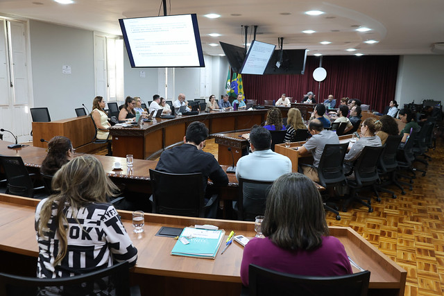 Além das pautas para votação, o Comitê Gestor também pode conferir os resultados de diferentes iniciativas em governança na UFC (Foto: Viktor Braga/UFC) Imagem: Visão ampla de uma reunião em formato de plenária, com diversas pessoas sentadas ao redor de uma grande mesa em U. Duas telas suspensas mostram a pauta da reunião. Participantes acompanham a apresentação, alguns fazendo anotações. No fundo da sala, estão as mesmas três pessoas da mesa principal vistas na primeira foto, com bandeiras do Brasil e de um estado posicionadas à esquerda.