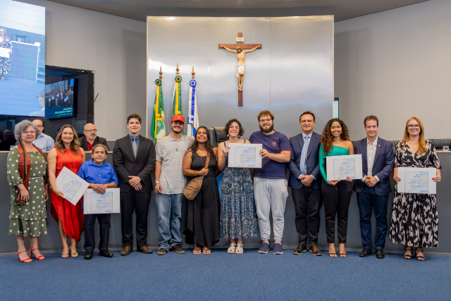 Homenageados posam ao lado da vice-reitora Diana Azevedo (vestido verde), do vereador Gabriel Aguiar (terno preto), do reitor Custódio Almeida (terno e gravata azuis) e do secretário de Juventude de Fortaleza, Júlio Brizzi (terno azul) (Foto: Guilherme Silva/UFC) Imagem: grupo de cerca de 14 pessoas, entre homens e mulheres de diferentes idades, tons de pele e estilos de roupa, posam em pé em um plenário. Algumas seguram certificados. Ao fundo, há um grande crucifixo fixado na parede, bandeiras do Brasil, do Ceará e de Fortaleza, e o espaço típico de uma câmara legislativa. Todos estão alinhados lado a lado, voltados para a câmera.