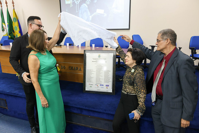 Imagem: foto em um palco de auditório. Quatro pessoas adultas participam do descerramento de uma placa comemorativa. Duas pessoas seguram um tecido branco sendo levantado, revelando uma placa emoldurada posicionada sobre um suporte à frente da mesa do palco. À esquerda, um homem e uma mulher observam o ato; à direita, outra mulher e um homem ajudam a puxar o tecido. Todos vestem roupas sociais. Ao fundo, aparecem cadeiras azuis e a mesa do palco, indicando um evento institucional ou solene.