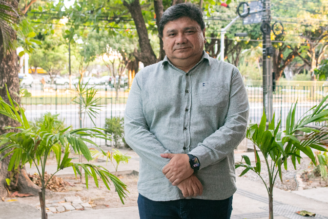 Imagem: homem branco, gordo, roupa social, cabelo preto com árvores ao fundo