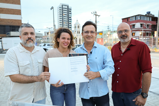 Imagem: De pé, da esquerda para a direita: João Wilson Damasceno (diretor do Instituto Iracema), Luisa Cela (secretária da Cultura), Custódio Almeida (reitor da UFC) e Sandro Gouveia (pró-reitor de Cultura da UFC). Eles sorriem e seguram o termo assinado nas mãos.