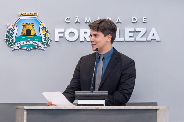 Autor do requerimento, o vereador Gabriel Aguiar (Psol) destacou, durante sua fala, números de infraestrutura, de serviços prestados, de recursos humanos e de conquistas da UFC, agradecendo a instituição pelo seu papel no desenvolvimento da cidade e do estado (Foto: Guilherme Silva/UFC) Imagem: homem jovem, de pele clara e cabelo castanho curto, veste terno escuro, camisa azul e gravata. Ele está em pé atrás de um púlpito, falando ao microfone e segurando um papel. Ao fundo, na parede clara, aparece o brasão e o nome “Câmara de Fortaleza”. O homem olha levemente para o lado, com expressão atenta e concentrada.