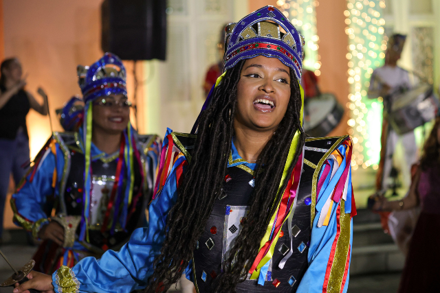 Imagem: Mulher com traje de reisado, fitas coloridas na cabeça e sorriso no rosto, em frente à Reitoria da UFC