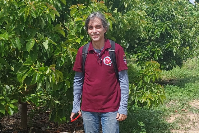Imagem: homem com cabelos lisos e parcialmente grisalhos posa para foto em um ambiente ao ar livre, cercado de verde e plantas. Ele veste camisa polo vermelha por cima de uma camisa de malha de manga longa, e calças jeans
