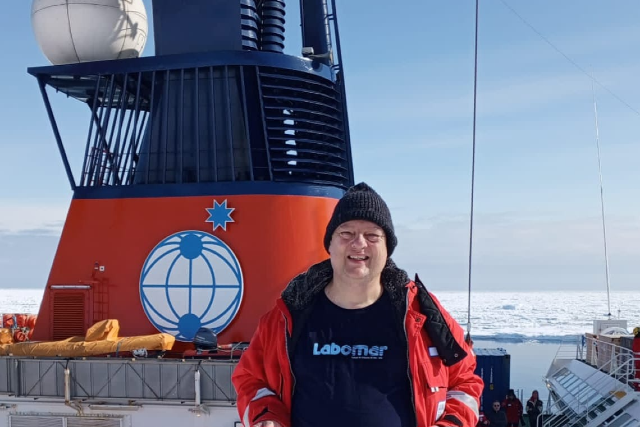 Imagem: Foto em ambiente polar. Em primeiro plano, um homem sorri para a câmera, vestindo gorro preto, jaqueta vermelha de frio intenso e camiseta azul-marinho com a palavra “Labomar” estampada. Ele está a bordo de um navio. Ao fundo, vê-se a grande chaminé do navio, nas cores azul e laranja, com um símbolo de globo estilizado e uma estrela, além de equipamentos e estruturas da embarcação. Atrás do navio, estende-se uma paisagem coberta por gelo e neve, típica da região da Antártida, sob céu claro.