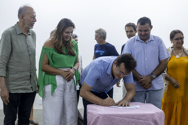 Imagem: Em uma sala interna, um homem inclinado para frente assina um documento sobre uma mesa coberta por uma toalha rosa-clara. Ao redor dele, várias pessoas estão em pé, observando o momento com atenção e expressões de interesse. O ambiente é claro e transmite clima formal e coletivo.