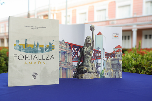Imagem: Foto do livro Fortaleza Amada sobre uma mesa; ao fundo, o prédio da Reitoria