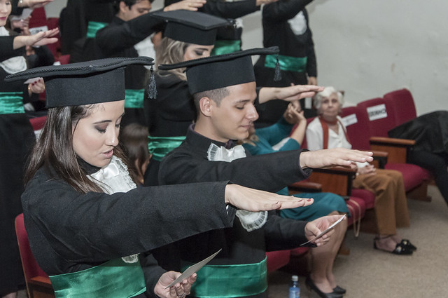 Imagem: Foto de cerimônia de colação de grau em auditório. Em primeiro plano, formandos e formandas sentados em cadeiras estofadas realizam o juramento profissional, com o braço direito estendido à frente. Eles vestem beca preta, faixa verde na cintura e capelo na cabeça, e seguram o texto do juramento nas mãos. Ao fundo, outros formandos repetem o gesto, enquanto algumas pessoas da plateia acompanham a solenidade sentadas nas fileiras do auditório.