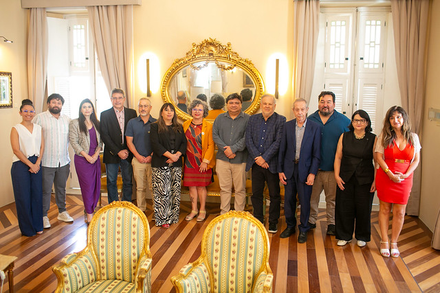 Imagem: Grupo de cerca de treze pessoas posa para foto institucional em uma sala elegante, com piso de madeira, poltronas claras e um grande espelho dourado ao fundo.