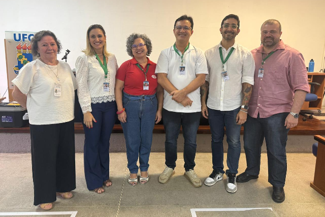 Imagem: Seis pessoas posam lado a lado para foto em uma sala institucional da UFC. Todas usam crachá e vestem roupas casuais ou sociais. Ao fundo, há uma mesa, cadeiras e um banner com a identidade visual da universidade.