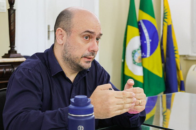 Imagem: Homem branco, de meia-idade, com cabeça parcialmente calva e barba curta, veste camisa social azul-escura e está sentado à mesa, em ambiente institucional. Ele gesticula com as mãos enquanto fala, demonstrando expressão atenta e concentrada.