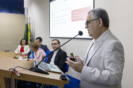 Imagem: Plano fechado do Dr. Fernando Barroso, chefe da Unidade de Hematologia e Hemoterapia do HUWC, falando ao microfone. Ao fundo, os integrantes da mesa solene o observam sentados.