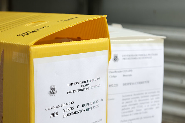 A doação do material para a associação cadastrada se dá por meio do Projeto Coleta Seletiva Cidadã (Foto: Viktor Braga/UFC Informa) Imagem: Fotografia de uma pilha alta de documentos impressos sobre uma mesa. As folhas brancas estão organizadas em vários blocos, alguns presos com clipes pretos. Entre as pilhas, há um envelope pardo parcialmente visível. Ao fundo, desfocado, aparecem prateleiras com pastas e fichários nas cores amarela, preta e branca, sugerindo ambiente de escritório ou arquivo.
