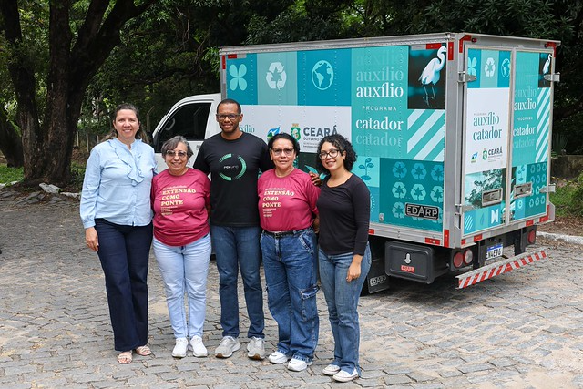 Marcela Teixeira (Memorial e CPAD), Margarida Sousa (secretária executiva da Prex), Josivan Soares Ferreira (Arquivista da SEAD/PROGEP e membro da CPAD), Maria Josineide Góis (Bibliotecária da PREX e membro da CPAD), Juliana Silva (Técnica de Laboratório/Química da Coordenadoria de Gestão de Resíduos da Secretaria do Meio Ambiente - SMAUFC) (Foto: Viktor Braga/UFC Informa) Imagem: Fotografia em ambiente externo, com árvores ao fundo e chão de paralelepípedo. Cinco pessoas posam lado a lado, sorrindo para a câmera. Duas mulheres usam camisetas vermelhas com a frase “Extensão como ponte”; as demais vestem roupas casuais, como camisa social clara e calça, camiseta preta e jeans. Ao lado do grupo, há um veículo utilitário adesivado nas cores verde e azul, com símbolos de reciclagem e textos como “auxílio catador” e referência ao Governo do Ceará.