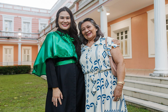 Imagem: Duas mulheres posam abraçadas em um jardim diante de um prédio histórico da universidade. A mulher à esquerda usa beca preta com capa verde e sorri para a câmera. A mulher à direita veste um vestido claro com estampa azul. Ao fundo aparecem gramado, colunas brancas e a fachada do edifício