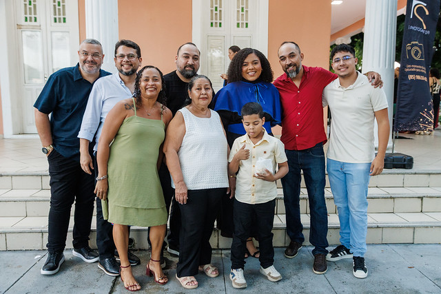 Imagem: Dez pessoas posam juntas em frente a um prédio claro, sorrindo para a câmera. Ao centro, está uma mulher negra de cabelo cacheado e veste talar azul