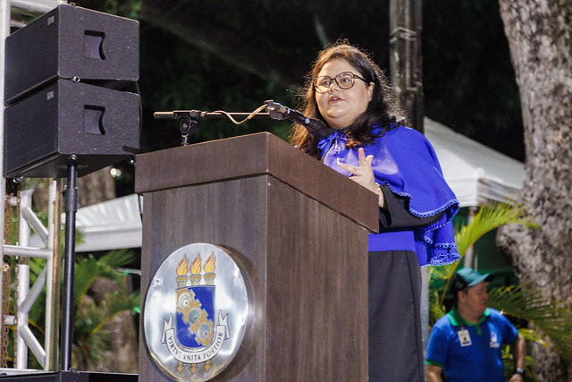 Imagem: Mulher jovem, de cabelos longos escuros e óculos, discursa atrás de um púlpito com o brasão da UFC. Ela veste beca preta e uma capa azul, e gesticula com uma das mãos enquanto fala ao microfone.