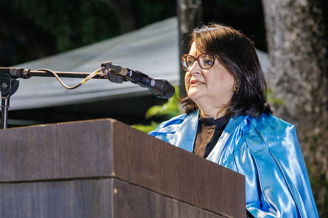 Imagem: Mulher de meia-idade, com cabelos escuros na altura dos ombros e óculos, fala ao microfone atrás de um púlpito de madeira com o brasão da UFC. Ela usa beca e uma capa azul sobre os ombros. A cena acontece à noite, em um espaço externo com iluminação artificial e vegetação ao fundo.