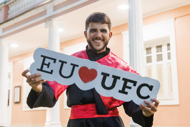 Imagem: Homem jovem, sorridente, veste beca preta com faixa vermelha e segura um letreiro branco escrito “EU ❤️ UFC”. Ele está em frente a um prédio histórico de paredes em tom salmão claro e colunas brancas