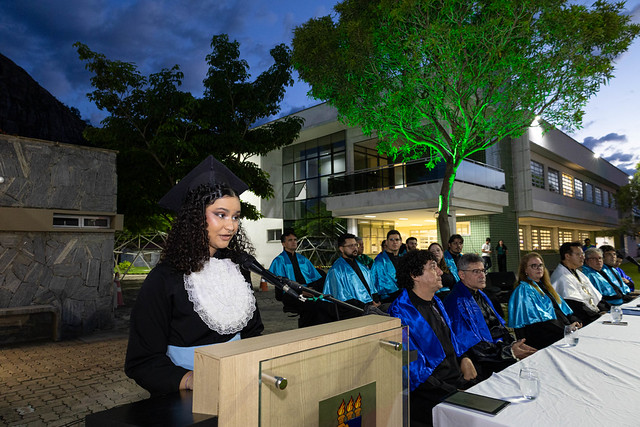 Imagem: Mulher jovem, de cabelos cacheados e escuros, veste beca preta com gola branca e fala ao microfone em um púlpito com o brasão da universidade. Ela aparece de perfil, olhando para a plateia. Atrás dela, em uma mesa longa coberta por toalha branca, estão sentadas várias autoridades acadêmicas usando capas azuis e douradas.