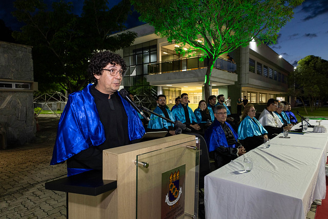 Imagem: Homem de cabelos cacheados e óculos fala ao microfone em um púlpito com o brasão da universidade. Ele usa roupa preta com capa azul brilhante. Atrás dele, outras autoridades acadêmicas, também com capas azuis e douradas, estão sentadas em uma mesa longa.