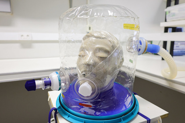 Imagem: Equipamento de laboratório utilizado para testes de máscaras respiratórias. No centro da imagem há uma cabeça de manequim dentro de uma câmara transparente de plástico, semelhante a um cilindro. O manequim está conectado a tubos e válvulas que simulam respiração. A base do equipamento é azul e o conjunto está sobre uma bancada de laboratório, com outros equipamentos