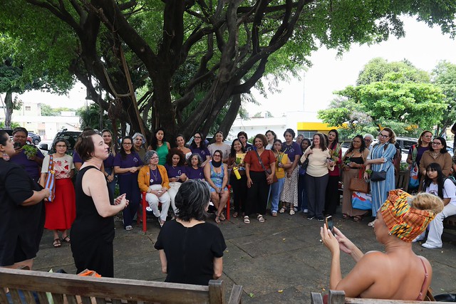 Imagem: Em área externa arborizada, um grupo numeroso de mulheres de diferentes idades e estilos de vestimenta se reúne em semicírculo sob uma grande árvore. Ao centro, uma mulher em pé fala para as demais, que a escutam com atenção