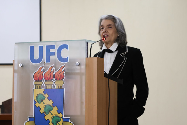 Imagem: Em auditório, uma mulher de cabelos grisalhos, vestindo blazer preto com detalhes claros, fala ao microfone em um púlpito com o brasão e a sigla UFC. Ela olha à frente com expressão firme, em momento de discurso.