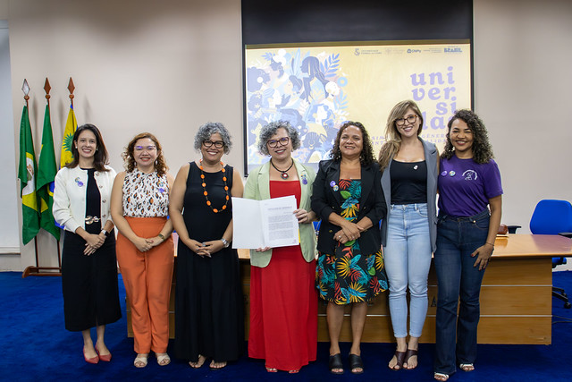 Imagem: Em sala institucional, sete mulheres posam lado a lado sorrindo para a câmera. Ao centro, uma delas segura um documento aberto. Ao fundo, há bandeiras e um painel com identidade visual do evento.
