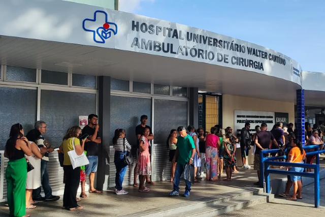 Imagem: Fotografia da entrada de uma unidade de saúde com a identificação “Hospital Universitário Walter Cantídio – Ambulatório de Cirurgia” na fachada. Em frente ao prédio, há uma fila de pessoas aguardando atendimento, distribuídas ao longo da calçada. Os indivíduos são de diferentes idades e vestem roupas casuais. À direita, há uma rampa com corrimão azul que dá acesso à entrada. O ambiente é externo, com iluminação natural e céu parcialmente visível ao fundo.