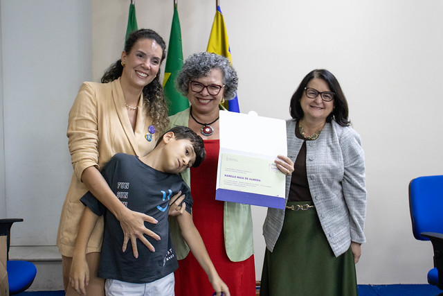 Imagem: Três mulheres e uma criança posam juntas sorrindo. Uma das mulheres segura um documento aberto voltado para a câmera. A criança, apoiada na lateral, inclina o corpo de forma descontraída. Ao fundo, bandeiras e cadeiras de auditório.