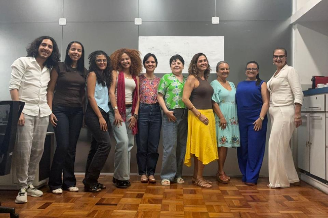 imagem: Fotografia em ambiente interno. Um grupo de dez pessoas posa lado a lado, sorrindo para a câmera. Há nove mulheres e um homem. Estão em pé sobre piso de madeira, em frente a uma parede cinza com um quadro branco ao centro. As pessoas vestem roupas variadas e coloridas, como calças, blusas, vestidos e sandálias. À direita da imagem, há um móvel branco com objetos apoiados. O clima é descontraído, de registro coletivo.
