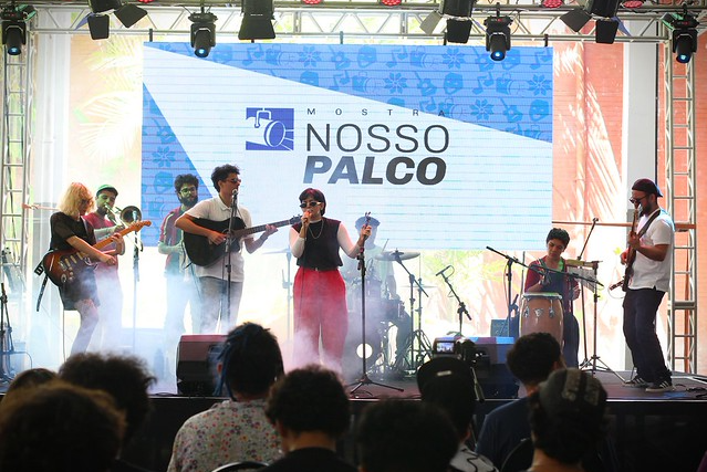 Imagem: Fotografia de apresentação musical em palco. Um grupo de artistas se apresenta diante de uma plateia. Ao centro, uma cantora segura um microfone, acompanhada por músicos com instrumentos como violão, guitarra, percussão e bateria. Ao fundo, um grande telão exibe o nome “Mostra Nosso Palco”. Há iluminação de palco com refletores coloridos e leve efeito de fumaça. Na parte inferior da imagem, aparecem as silhuetas do público assistindo ao show.