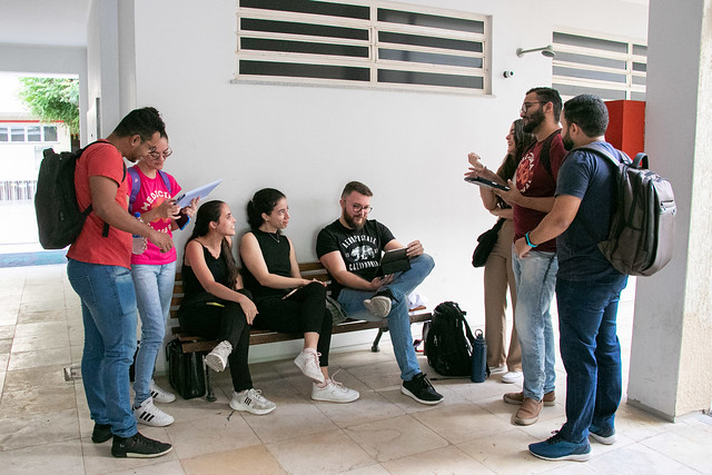 Imagem: Fotografia em corredor interno de prédio universitário. Um grupo de oito estudantes conversa e realiza atividades acadêmicas. Três pessoas estão sentadas em um banco, usando cadernos e computador portátil, enquanto as demais permanecem em pé, segurando folhas e conversando entre si. Os estudantes vestem roupas casuais e carregam mochilas. O ambiente possui paredes brancas, piso claro e iluminação natural.