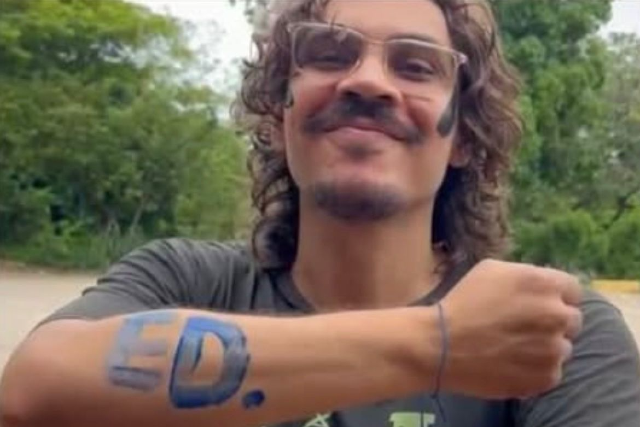 Imagem: Homem sorrindo mostra o antebraço com a palavra “ED.” pintada em azul. Fundo com árvores ao ar livre.