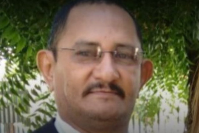 Imagem: Homem negro, com cabelo curto e escuro, usando óculos de armação fina e bigode. Ele veste roupa social e está em ambiente externo, com árvores ao fundo. A expressão é neutra e o rosto aparece em close.