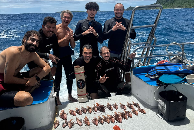 Imagem: Sete mergulhadores posam sobre uma embarcação em alto-mar, sob céu parcialmente nublado. O grupo, formado por homens, veste roupas de mergulho pretas; alguns estão descalços e outros usam parte do equipamento. Todos sorriem para a câmera e fazem gestos com as mãos.  À frente deles, sobre o piso branco do barco, estão dispostos vários peixes-leão alinhados em fileiras, resultado da ação de manejo da espécie invasora. Também aparecem equipamentos de mergulho, como nadadeiras azuis, cilindro e baldes. Ao fundo, o mar azul se estende até uma faixa de vegetação em terra firme, visível no lado direito da imagem.