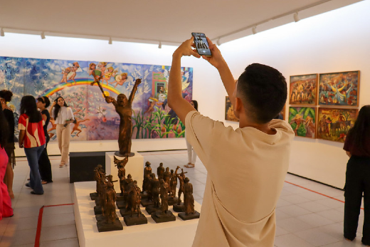 Imagem: A imagem mostra o interior de uma galeria do Museu de Arte da UFC, com visitantes observando uma exposição de pinturas e esculturas. Em primeiro plano, aparece um homem de costas, usando camisa clara, segurando um celular acima da cabeça para fotografar ou filmar as obras expostas. À frente dele, sobre uma base branca, há diversas esculturas pequenas em tom escuro, organizadas em conjunto, além de uma escultura vertical maior ao centro.  Nas paredes ao fundo, destacam-se pinturas coloridas e expressivas, com cenas figurativas e elementos humanos. À esquerda, um grande quadro em tons de azul, lilás e rosa ocupa boa parte da parede. À direita, há uma sequência de telas menores com cores quentes e composição vibrante. Outros visitantes circulam pelo espaço, observando as obras e conversando. O ambiente é amplo, iluminado por luzes direcionadas no teto, com paredes brancas e atmosfera contemplativa típica de um museu de arte contemporânea.