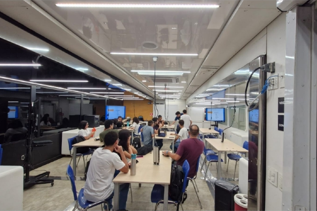 Imagem: imagem de dentro de uma sala de aula com mesas, cadeiras, estudantes de costas olhando pra uma lousa e sentados ao redor de mesas. Local com janelas de vidros.