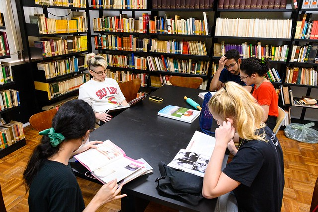 Imagem: Sala de biblioteca com estantes altas repletas de livros organizados ao fundo e nas laterais. No centro, uma mesa retangular preta reúne cinco estudantes sentados, concentrados na leitura e consulta de materiais.  Uma estudante de cabelos escuros presos por um laço verde folheia um livro aberto. À frente, outra estudante loira consulta um livro, apoiando uma das mãos no rosto. Ao lado, dois estudantes observam o mesmo material, enquanto uma estudante de cabelos claros, sentada ao fundo, lê um livro com atenção. Sobre a mesa, há livros, cadernos, estojo e garrafas. O ambiente é iluminado, silencioso e organizado, sugerindo momento de estudo coletivo e pesquisa acadêmica