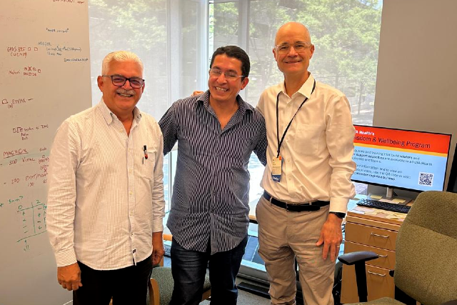 Imagem: Três homens adultos posam sorrindo em um escritório iluminado. À esquerda, um homem de cabelos brancos e óculos veste camisa social clara. No centro, um homem de camisa azul listrada abraça os colegas pelos ombros. À direita, um homem careca usa camisa branca social e crachá pendurado no pescoço.  Ao fundo, há uma grande janela com vista para árvores, uma mesa de escritório com computador e cadeira, além de um quadro branco com anotações e fórmulas escritas em diferentes cores. Na tela do computador, aparece uma apresentação relacionada a saúde e bem-estar. O ambiente transmite contexto acadêmico, científico ou institucional.