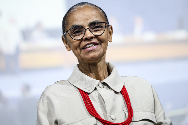 Imagem: Marina Silva em um retrato de meio corpo, posicionada de frente para a câmera e com uma leve inclinação da cabeça. Ela sorri suavemente, transmitindo uma expressão acolhedora e serena.  A pessoa usa óculos de armação fina e tem os cabelos grisalhos presos para trás de forma discreta. Está vestida com uma camisa de tom bege claro, de mangas compridas, com gola estruturada e botões frontais. Como acessório de destaque, usa um colar longo composto por várias voltas de contas vermelhas, que contrastam com a neutralidade da roupa.